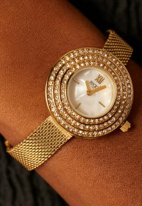 Gold mesh wristwatch with circular face, white mother-of-pearl dial, and sparkling crystal accents around the bezel. Features gold hour markers.