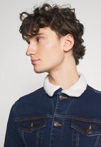 Jeans jakna v temno modri barvi z belim flis ovratnikom, ki ima gumbne zapore in dve žepni torbici na prsih. Obrnjen hem in vidne šivane podrobnosti.