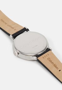 Montre Calvin Klein avec un boîtier en acier inoxydable argenté, un bracelet en cuir noir et un logo gravé au dos poli. Comprend une couronne.