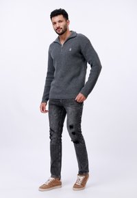 Grauer, gestrickter Pullover mit Reißverschlusskragen und Logodetails, kombiniert mit ausgebeulten schwarzen Jeans und hellbraunen, lässigen Turnschuhen.
