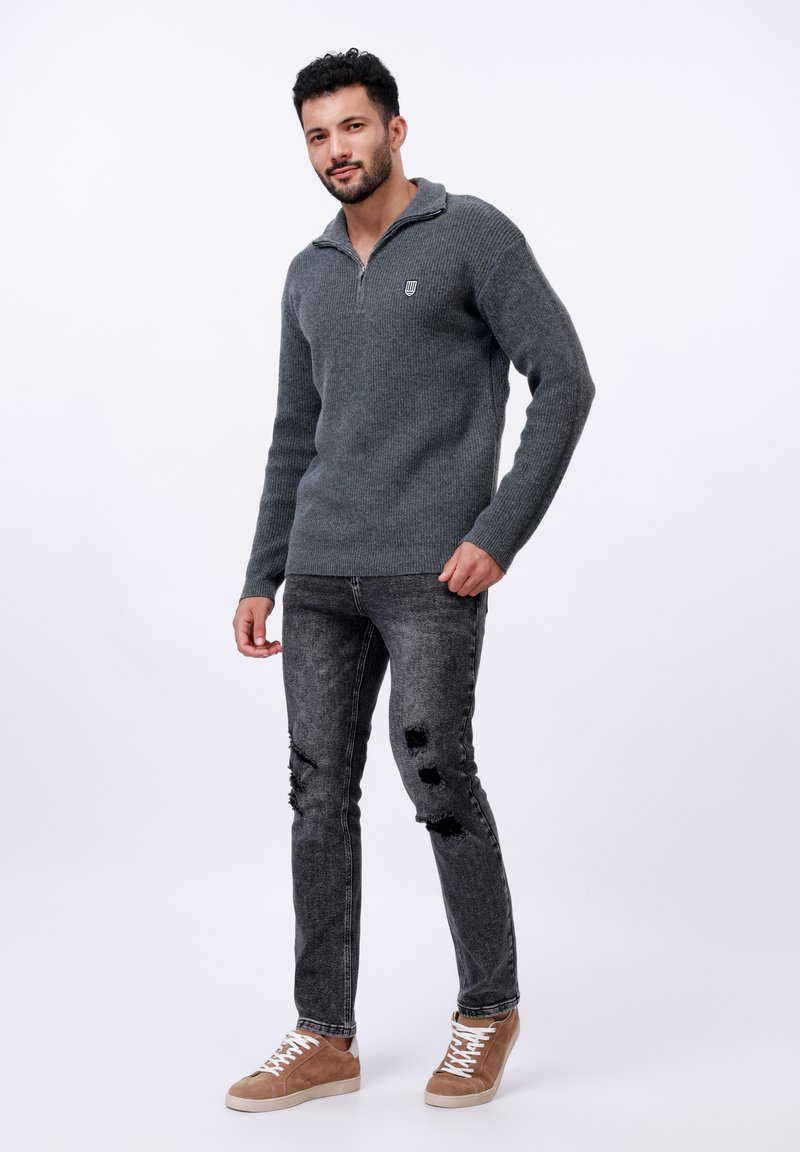 Grauer, gestrickter Pullover mit Reißverschlusskragen und Logodetails, kombiniert mit ausgebeulten schwarzen Jeans und hellbraunen, lässigen Turnschuhen.