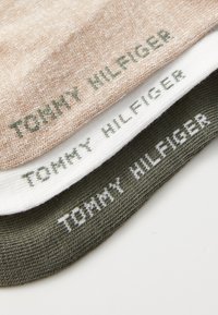 Tre foldede sokker i beige, hvid og mørkegrøn, hver med "Tommy Hilfiger" strikket på kanten i kontrastfarve.
