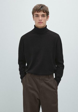 Pull en col roulé noir en matériau doux, manches longues, associé à un pantalon marron, sur fond pâle.