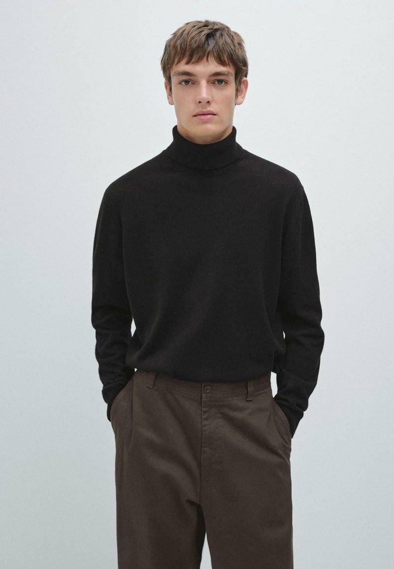 Maglione nero a collo alto realizzato in materiale morbido, maniche lunghe, abbinato a pantaloni marroni, sullo sfondo chiaro.