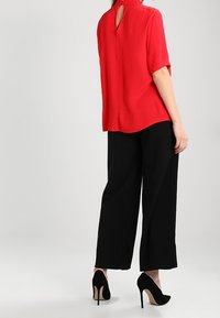 Blusa rossa a maniche corte con apertura a chiave sul retro, abbinata a pantaloni neri a gamba larga e scarpe nere con tacco alto, caratterizzata da una texture liscia.