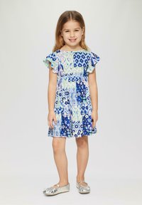 Blau gemustertes Kleid mit schwingenden Ärmeln, das florale und geometrische Designs aufweist. Kombiniert mit glänzenden silbernen Ballerinas. Hautfarbe hell.