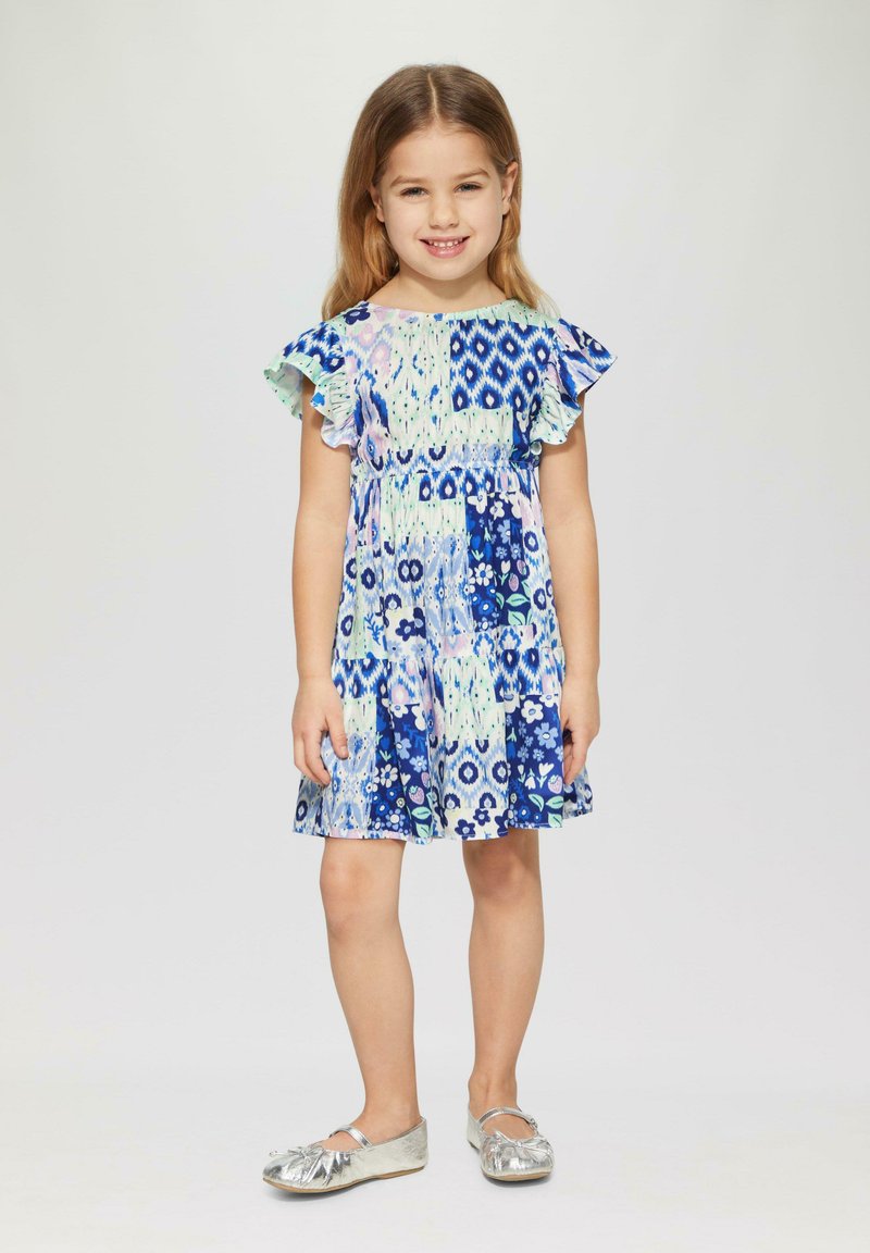 Blau gemustertes Kleid mit schwingenden Ärmeln, das florale und geometrische Designs aufweist. Kombiniert mit glänzenden silbernen Ballerinas. Hautfarbe hell.