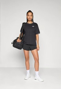 Melna Puma T-krekls ar īsām piedurknēm un grafisku logo, kombinēts ar melniem šortiem. Modelis rokās tur melnu sporta somu, valkā baltus sporta apavus un zeķes.