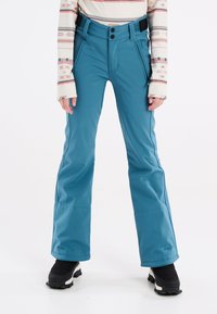 Pantaloni da sci impermeabili color turchese con gamba svasata, tasche con zip e chiusura a due bottoni. Tessuto testurizzato con cuciture rinforzate; abbinati a stivali neri.