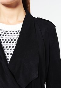 Cappotto avvolgente in camoscio nero con colletto a scialle e spalline sulle spalle, indossato sopra un maglione bianco con motivi geometrici neri.