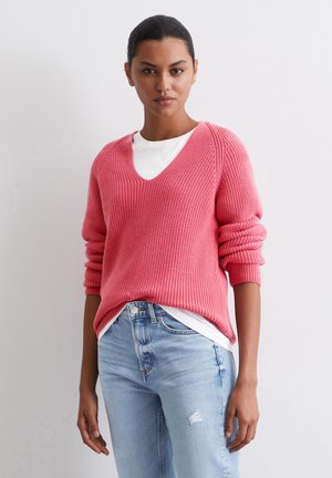 Rosa Strickpullover mit V-Ausschnitt, geripptem Muster und überschnittenen Schultern, kombiniert mit einem weißen Unterhemd und hellblauen, distressed Jeans.