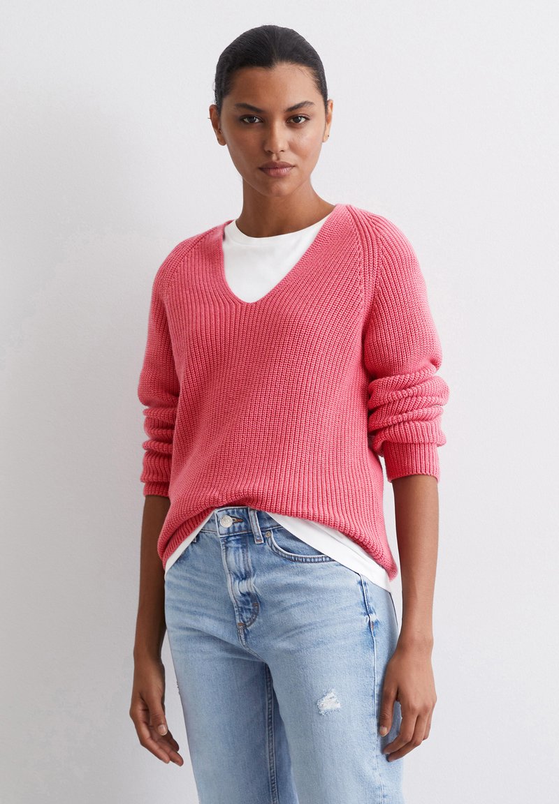 Roze gebreide trui met een V-hals, geribbeld patroon en verlaagde schouders, gecombineerd met een witte ondertop en lichtblauwe versleten jeans.