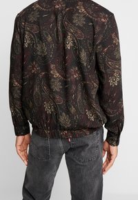 Man in een donker paisley-patroon overhemd met lange mouwen, in zijn zwarte vervaagde jeans gestoken, gezien vanuit de achterkant.