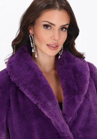 Vrouw met lang bruin haar, gekleed in sprankelende parel- en rhinestone-druppeloorbellen en een levendige paarse nep-bontjas.