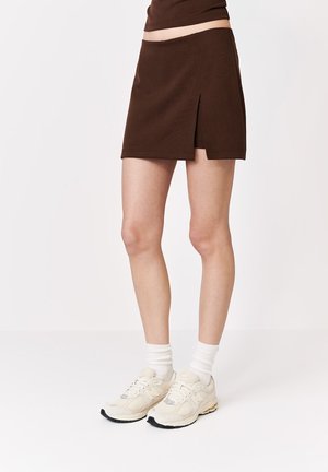 Osoba nosząca brązową mini spódniczkę z bocznym rozcięciem, białe skarpety i beżowe trampki stoi na białej podłodze na jednolitym tle.