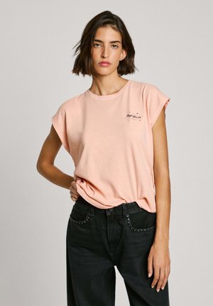 T-shirt di cotone color pesco con maniche arrotolate, caratterizzato da un piccolo design grafico nero sul petto e una vestibilità rilassata. Abbinato a jeans neri.