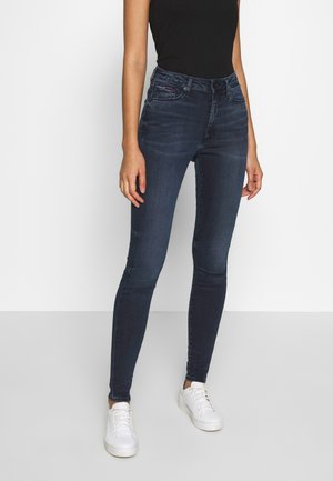 Jean skinny bleu foncé en denim, avec une taille haute, un léger délavage, et des poches avant et arrière. Associé à des baskets blanches.