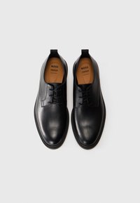 O pereche de pantofi de dress pentru bărbați, din piele neagră, cu șireturi, având interiorul de culoare bej, etichetați "BOSS Premium Comfort", pe un fundal alb.