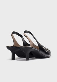Tacones slingback de piel negra con punta redonda, tacón medio cuadrado y tachuelas metálicas decorativas en la parte delantera. Textura suave, diseño minimalista.