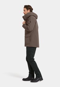 Bruine waterdichte parka met een hoge kraag en capuchon, voorzien van zijzakken en zwarte accenten. Gecombineerd met zwarte broeken en botten.