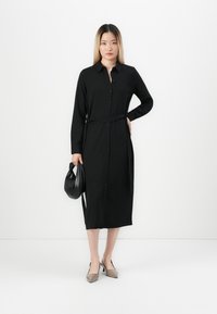 Woman wearing a long black buttoned dress with a belt, holding a black bag, standing against a plain white background.