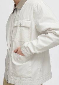 Persoon in een lichtbeige ritsjack met een klepborstzak en een hand in een lagere voorzak.