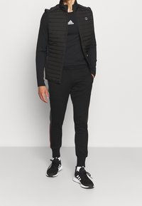 Schwarze, gepolsterte Weste über einem schwarzen Langarmshirt, kombiniert mit schwarzen Jogginghosen mit farblichen Akzenten und schwarzen Sportschuhen.