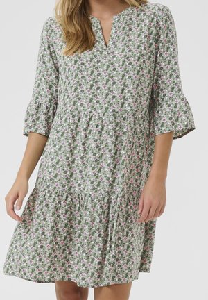Frau trägt ein grünes, geblümtes knielanges Kleid mit 3/4 Glockenärmeln und einem kleinen V-Ausschnitt.