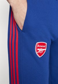 Modré sportovní kalhoty vyrobené z měkké tkaniny, s červenými bočními pruhy a vyšitým logem Arsenalu na stehně. Součástí jsou kapsy.