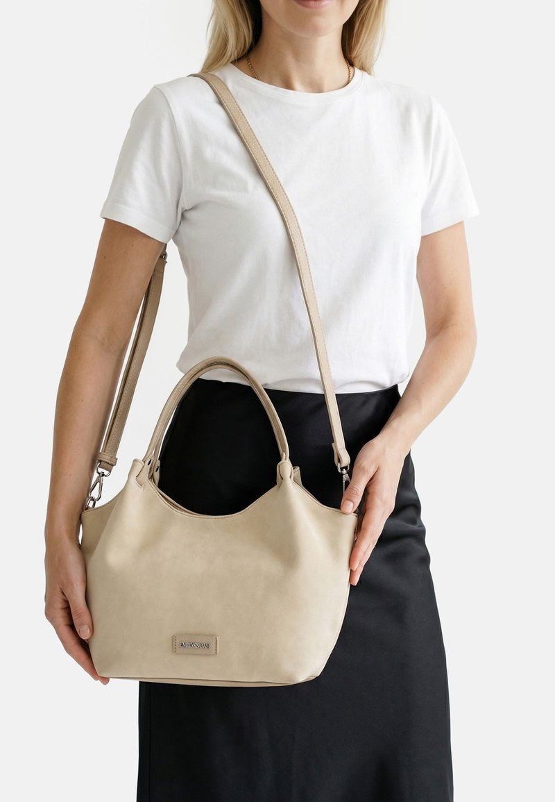 Mujer con camiseta blanca y falda negra sostiene un bolso beige por ambas asas y la correa cruzada, sobre un fondo blanco.