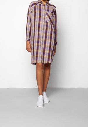 Femme portant une robe chemise longue jusqu’aux genoux, manches longues, à carreaux violets et marron, avec des baskets blanches, debout devant un fond uni.