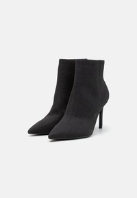 Botas de tobillo de punto negro con punteras puntiagudas y tacones de aguja. La tela texturizada tiene un brillo sutil y forma una silueta ajustada.