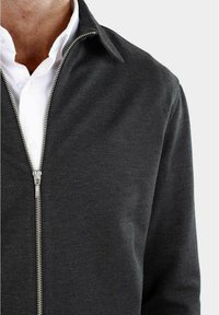 Donkergrijze ritsjack met een gladde textuur en een gestructureerde kraag. Voorzien van een zilveren rits en gedragen over een wit shirt.