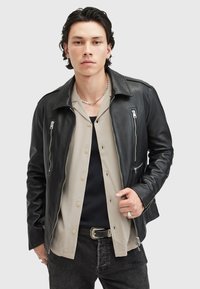 Chaqueta de cuero negra con un diseño clásico, que presenta cremalleras en el pecho y los laterales, combinada con una camisa beige sobre una camiseta negra.