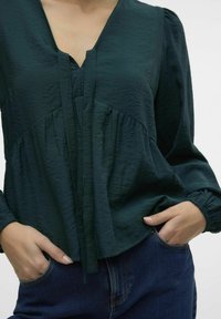 Mørkegrøn, struktureret bluse med V-udskæring, plissé-detaljer og lange pufærmer. Sæt med mørk denim med lomme-sømme.