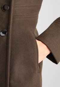 Abrigo de lana marrón con un diseño estructurado, detalles de costura horizontal, un botón negro único y un bolsillo con una mano dentro.