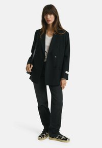 Blazer noir à double boutonnage avec deux boutons, porté sur un top blanc et assorti à un jean foncé et des chaussures à enfiler à motifs.