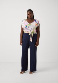 Blouse à motifs floraux avec un col arrondi et des manches courtes, associée à un pantalon large bleu marine. La blouse présente un détail de taille nouée et des couleurs vives.