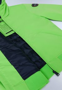 Giacca verde neon con un esterno liscio, dotata di una foderatura trapuntata blu navy, polsini a costine e un'etichetta con le istruzioni per la cura.