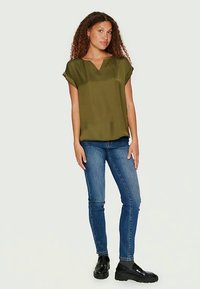 Olivno zelena bluzica s kratkimi rokavi in V-izrezom, teksturirana tkanina, ohlapen kroj, v kombinaciji z modrimi skinny kavbojkami in črnimi platform čevlji.