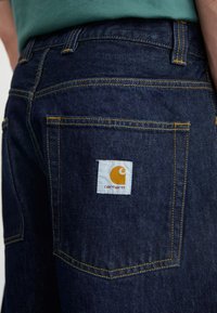 Temno modri kavbojki z rumenim šivom, ki imata zadnji žep in oranžno-belo oznako Carhartt.