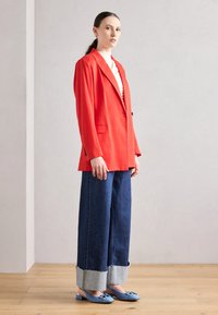 Červený blejzer so tailored dizajnom, skombinovaný s širokými tmavými džínami s rovnými okrajmi. Modré slip-on topánky dokončujú vzhľad.