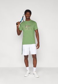 Πράσινο κοντομάνικο μπλουζάκι με μαύρο γραφικό σχέδιο, σε συνδυασμό με λευκά σορτς με πράσινες πλευρικές ρίγες και λευκά αθλητικά παπούτσια.