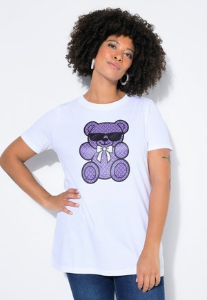 Kobieta z kręconymi włosami, nosząca kolczyki koła oraz białą koszulkę z fioletowym misiem z okularami przeciwsłonecznymi i muszką.