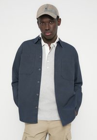 Νέος άνδρας που φοράει μπεζ καπέλο, μπλε σκούρο πουκάμισο, ανοιχτό γκρι πόλο και μπεζ παντελόνια εργασίας, στέκεται μπροστά σε απλό λευκό φόντο.