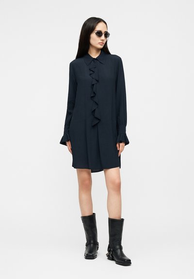 Γυναίκα με μακριά σκούρα μαλλιά, που φοράει μαύρα γυαλιά ηλίου, μαύρο φόρεμα με μακριά μανίκια και βολάν μπροστά, καθώς και μαύρα μποτάκια έως τη μέση της γάμπας, στέκεται μπροστά σε λευκό φόντο.