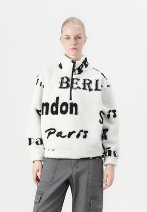 White fleece pullover with high collar featuring black text, including city names. Textured fabric, zip closure, relaxed fit. Gray cargo pants.