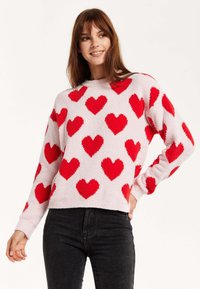 Light pink sweater with fluffy red heart patterns. Crew neckline, long sleeves, and a relaxed fit. Paired with black jeans.