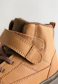 Helle Leder-Hochschuh mit einem strukturierten Stoffriemen, kontrastierenden Nähten und einer Gummisohle für besseren Halt.