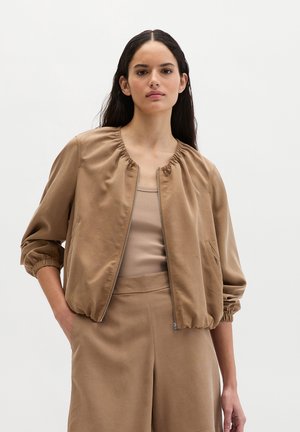 Frau mit langen dunklen Haaren, die eine beige Bomberjacke über einem passenden Top und einer Hose trägt, stehend vor einem schlichten, hellen Hintergrund.